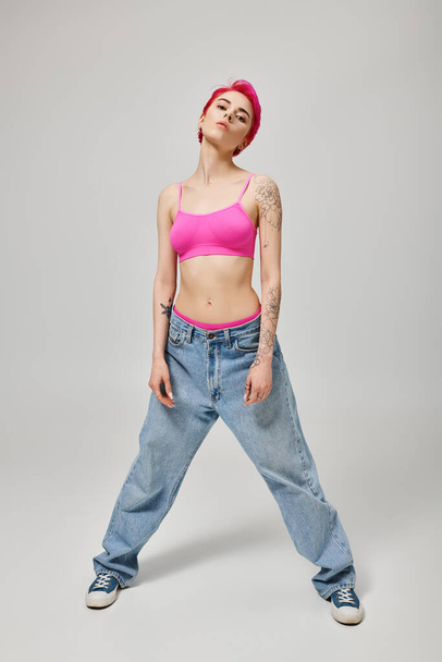 full length, confident woman with pink short hair posing in crop top and jeans on grey backdrop - Fotoğraf, Görsel