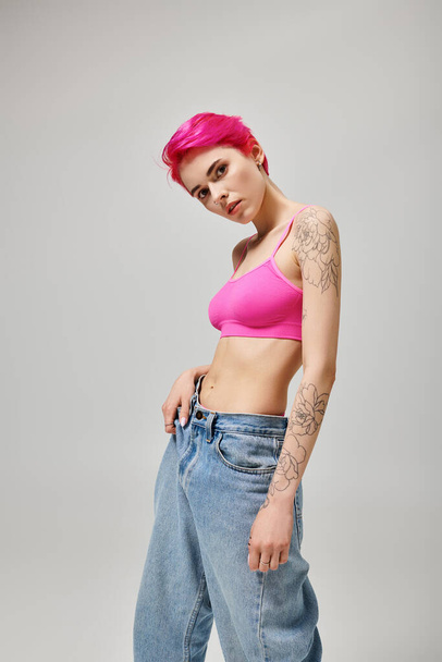 молода впевнена жінка з рожевим коротким волоссям позує у верхній частині культури і джинси на сірому фоні - Фото, зображення