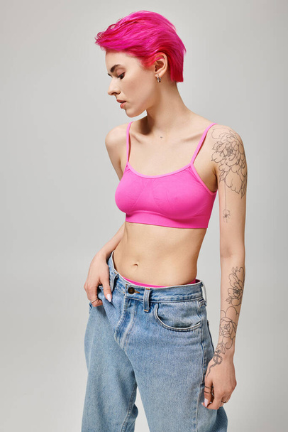 magabiztos és tetovált fiatal nő rózsaszín haj pózol a termés felső és farmer szürke háttér - Fotó, kép