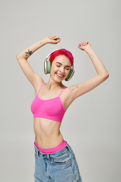 χαρούμενη και τρυπημένη γυναίκα με ροζ μαλλιά ακούγοντας μουσική σε ασύρματα ακουστικά σε γκρι φόντο - Φωτογραφία, εικόνα