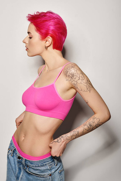 グレーの背景にクロップトップとジーンズでポーズピンクの髪のタトゥーされた若い女性の側面ビュー - 写真・画像