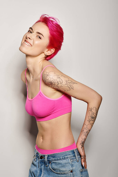 tattooed and expressive woman with pink hair posing with hands in pockets of jeans on grey - Photo, Image