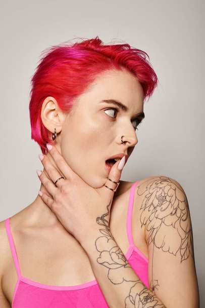 mujer tatuada y perforada con pelo rosa y anillos en los dedos mirando hacia otro lado sobre fondo gris - Foto, Imagen