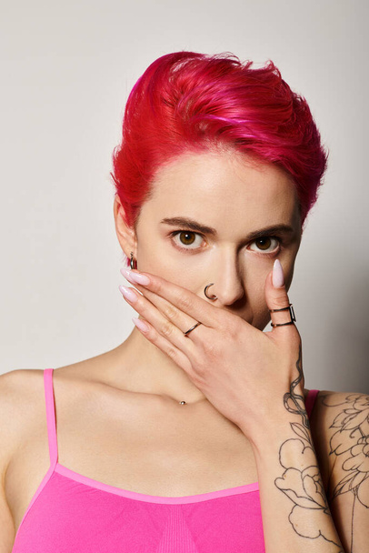 ausdrucksstarke junge Frau mit rosa Haaren, die den Mund bedeckt und vor grauem Hintergrund in die Kamera blickt - Foto, Bild