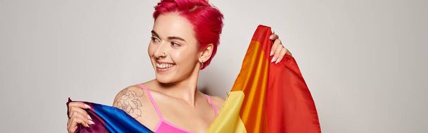 retrato de activista mujer complacida con el pelo rosa posando con la bandera del arco iris lgbt en gris, bandera - Foto, Imagen