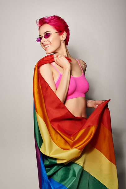 young and happy female activist with pink hair and sunglasses posing with lgbt rainbow flag on grey - Photo, Image