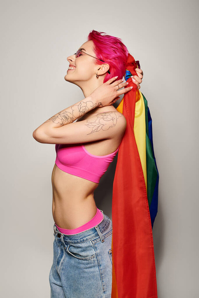 young and jolly woman with pink hair and sunglasses posing with lgbt rainbow flag on grey backdrop - Photo, Image