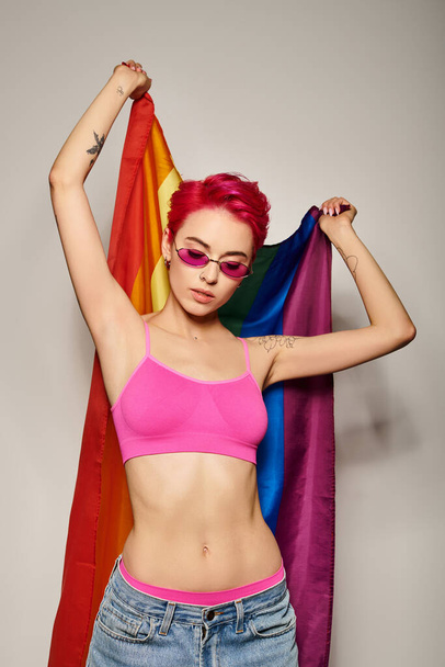 mujer joven tatuada con pelo rosa y gafas de sol posando con bandera de arco iris lgbt sobre fondo gris - Foto, Imagen