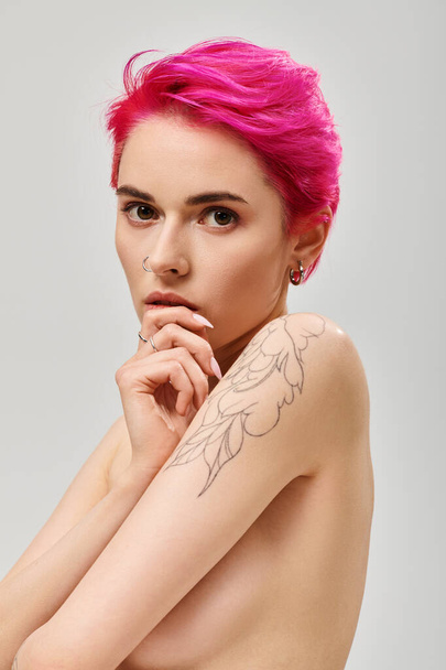 retrato de mujer joven tatuada y en topless con el pelo rosa cubriendo los pechos en el fondo gris - Foto, Imagen