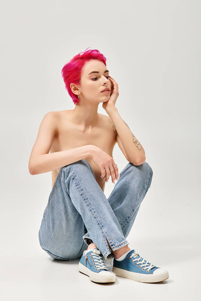tatuoitu ja yläosattomissa nainen vaaleanpunaiset hiukset istuu farkut harmaalla pohjalla, rento aiheuttaa - Valokuva, kuva
