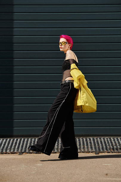 黄色のサングラスと屋外の都市の通りを歩くスタイリッシュな服のピンクの髪の若い女性 - 写真・画像