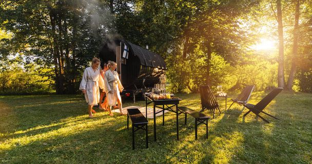 Pessoas relaxadas em roupões vêm de uma cabine de sauna em barril de madeira em um exuberante parque verde ao pôr-do-sol. São relaxados, rindo e desfrutar das férias enquanto relaxa na cabine de sauna finlandesa. - Foto, Imagem