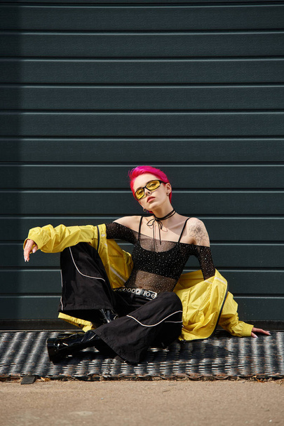 pink haired young woman in yellow sunglasses and stylish attire sitting on urban street outdoors - Photo, Image