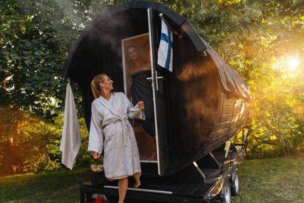 Pessoas em roupões de banho em frente a um barril de sauna finlandês móvel com vapor ao seu redor - Foto, Imagem
