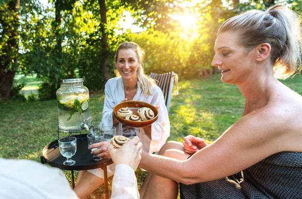 Mujeres en albornoces disfrutando de bocadillos de canela y bebidas en un jardín, con una sauna y dispensador de bebidas en el fondo - Foto, Imagen
