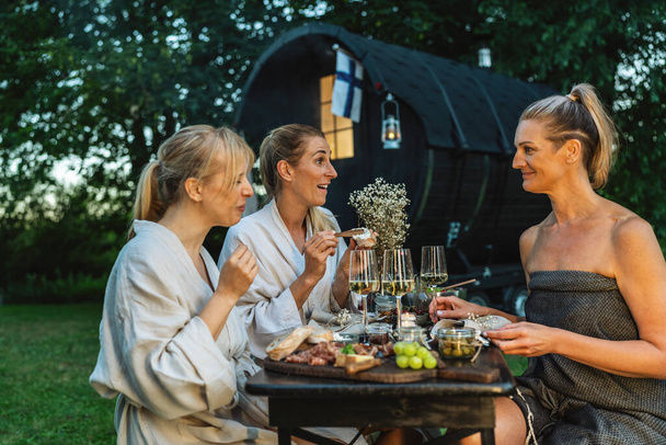 Amigos en albornoces disfrutando de una comida y vino cerca de una sauna finlandesa al aire libre - Foto, imagen