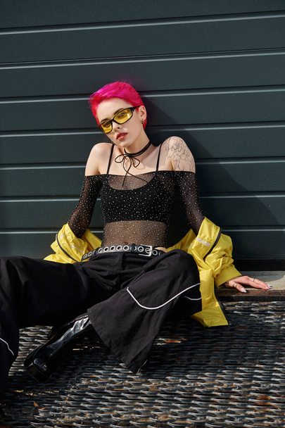 hübsche Frau mit pinkfarbenen Haaren und Tätowierung posiert in Sonnenbrille und trendiger Streetwear auf urbaner Straße - Foto, Bild