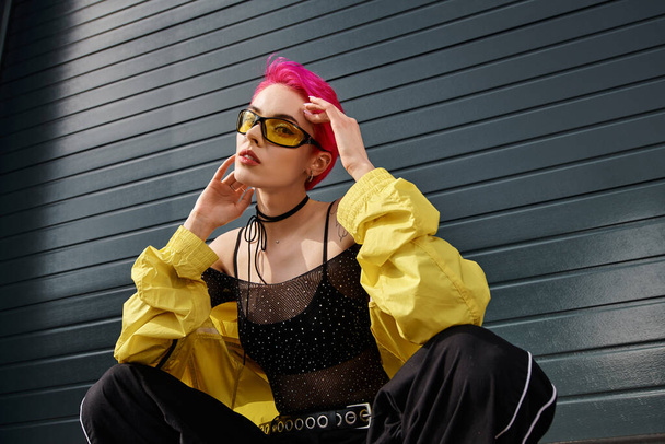 junge schicke Frau mit rosa Haaren und Tätowierung posiert mit Sonnenbrille und trendiger Streetwear auf der Straße - Foto, Bild