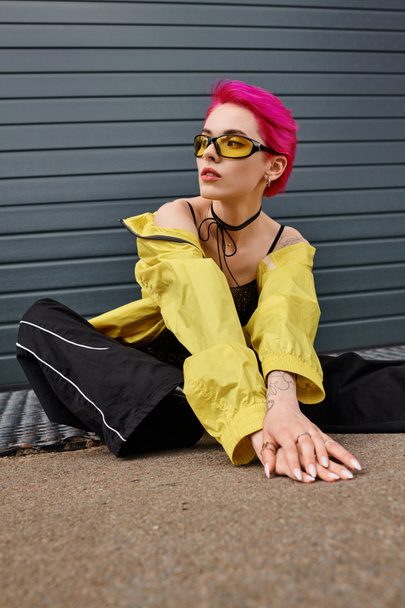 tetovált és rózsaszín hajú fiatal nő sárga napszemüvegben és elegáns öltözékben pózol a városi utcán - Fotó, kép