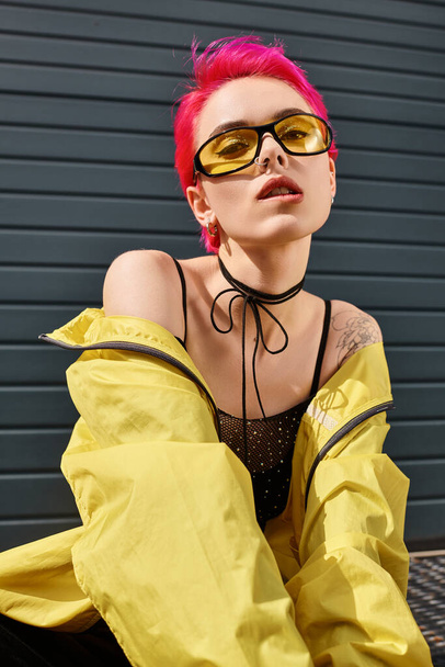 mujer joven de pelo rosa en gafas de sol amarillas y elegante traje mordiendo el labio y mirando a la cámara - Foto, imagen