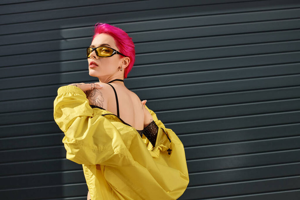 rózsaszín hajú fiatal nő sárga napszemüveg és divatos ruhát pózol és nézi a kamera - Fotó, kép