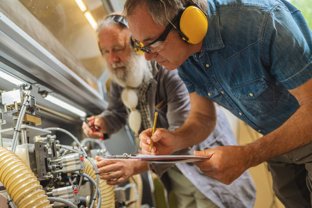 Due uomini con paraorecchie che lavorano su macchinari in una falegnameria - Foto, immagini