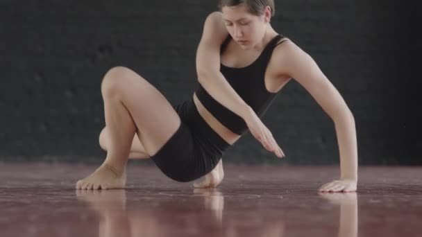 Joven talentosa mujer caucásica descalza usando top negro ajustado y pantalones cortos bailando elegante danza contemporánea en el suelo de parquet rojo en el estudio - Metraje, vídeo