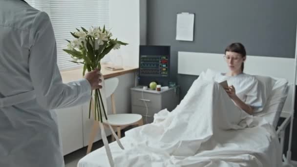Tracking over shoulder shot of female Caucasian patient lying in hospital bed receiving flowers from her unrecognizable doctor and reading note from bouquet - Footage, Video