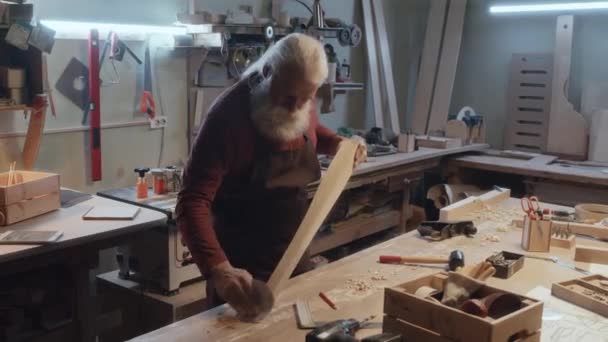 Μέτριο πλήρες πλάνο του παλαιού Καυκάσιου ξυλουργού με θαμνώδη γκρίζα γενειάδα λειαίνοντας ξύλινο στέλεχος με γυαλόχαρτο - Πλάνα, βίντεο