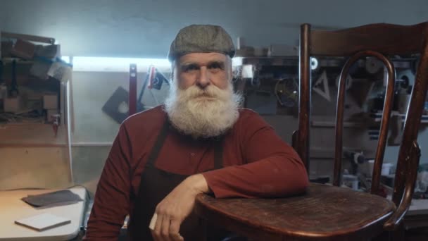 Střední portrét postarší bělošský tesař v šedém klobouku a zástěře opírající se o dřevěnou židli při pohledu na fotoaparát s úsměvem - Záběry, video