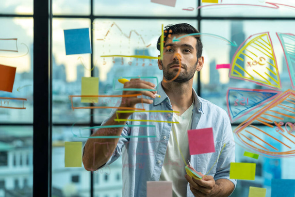 Portrait of young handsome businessman writing marketing idea by using colorful marker on glass wall with graph and mind map. Manager sharing idea or financial strategy. Business plan. Tracery. - Photo, Image