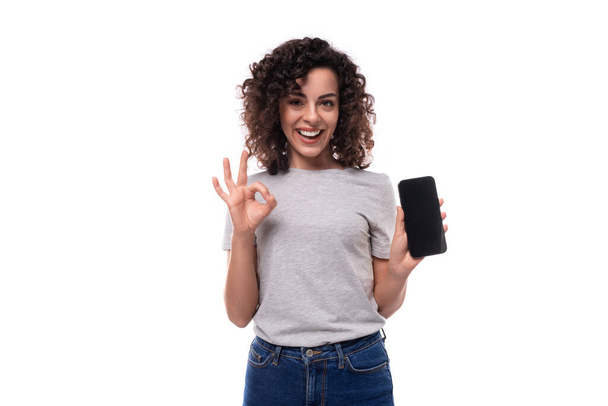 confident young curly brunette woman in gray t-shirt showing smartphone screen with mockup isolated on white background with copy space. - Photo, Image