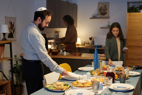 Widok z boku brodatego Żyda w czaszce służącego do stołu na obiad podczas przygotowań do obchodów Chanuki z rodziną w domu - Zdjęcie, obraz