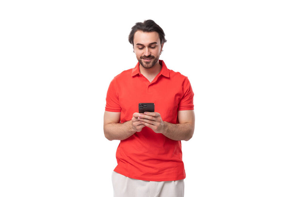 joven guapo europeo morena hombre con una barba vestida con una camiseta roja con una maqueta utiliza un teléfono móvil. - Foto, Imagen