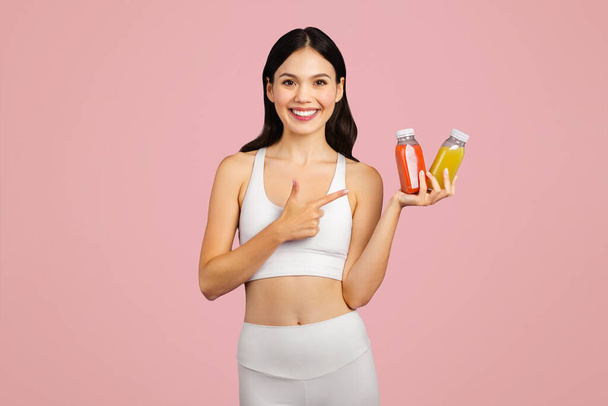 Positive caucasian lady in sportswear holding and pointing at jars of juice or smoothies isolated on pink background. Recommendation vitamins for sports, diet and health drink, body care - Photo, Image