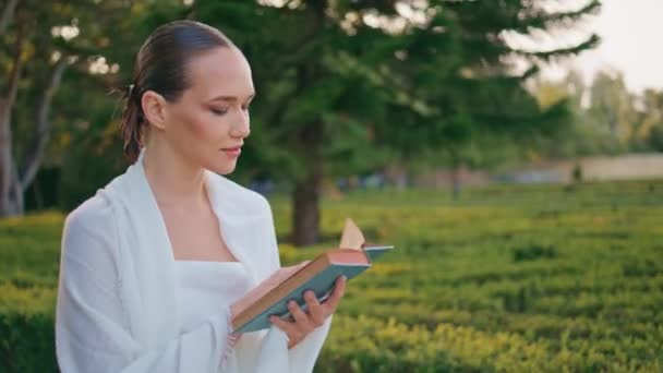 Romantic lady reading novel on go at beautiful empty park close up. Attractive dreamy woman focused on interesting literature walking green garden alone. Gentle young girl turning pages of book. - Footage, Video