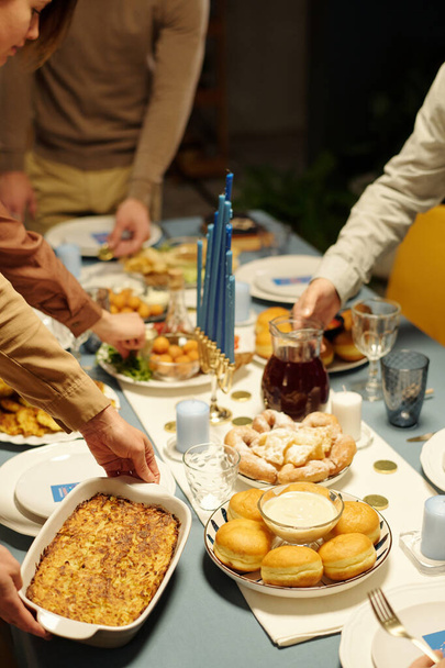 Ręce żydowskich członków rodziny umieszczając sztućce z apetycznym domowym jedzeniem na stole serwowane na obiad Chanuka - Zdjęcie, obraz
