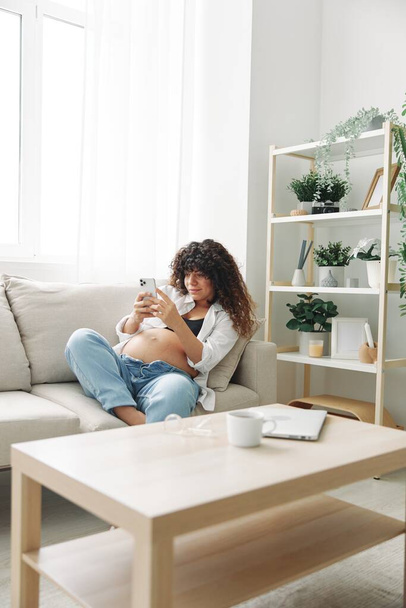 A blogueira grávida senta-se no sofá em casa e tira fotos de si mesma no telefone, selfie e videochamada, consulta com o médico on-line, gerenciamento de gravidez. Foto de alta qualidade - Foto, Imagem