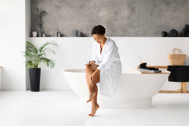 Beautiful black female applying moisturising cream on legs while sitting in luxury bathroom interior, happy african american woman in white bathrobe making beauty treatments at home, copy space - Foto, Imagen
