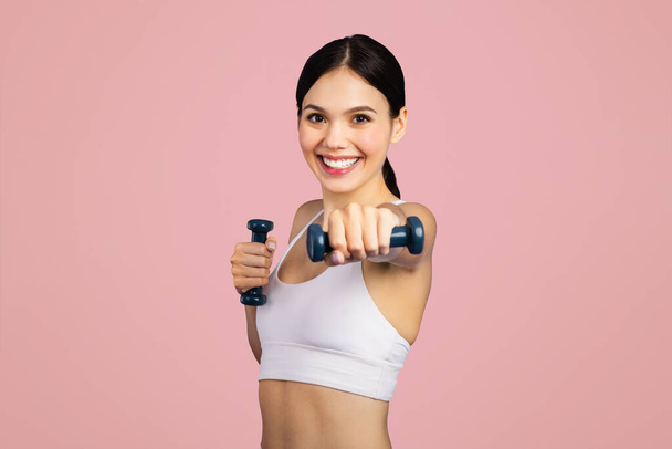 Aktivní běloška ve sportovním oblečení dělá paže cvičení s činkami na růžovém pozadí studia. Cvičení, péče o tělo s vybavením, silový trénink - Fotografie, Obrázek