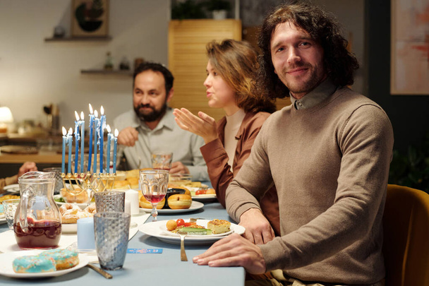 Onnellinen nuori juutalainen mies katselee kameraa istuessaan pöydän ääressä tarjoillen kotitekoista ruokaa ja juomia vaimoaan ja isäänsä vastaan. - Valokuva, kuva