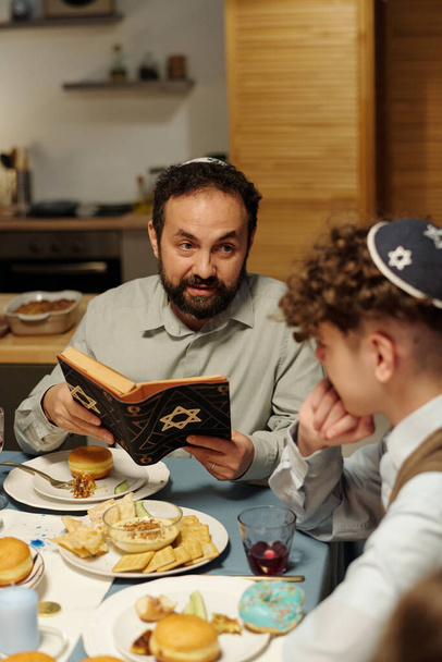 Focus on bearded mature man in kippah looking at his pre-teen son while explaining him text from Torah by festive Hanukkah dinner - Photo, Image