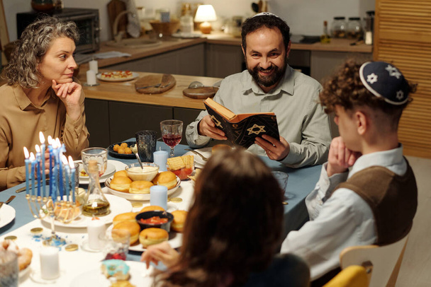 Щасливий бородатий чоловік з відкритим Писанням сидить перед своєю сім'єю і дивиться на дочку під час обговорення тексту зі Старого Заповіту - Фото, зображення