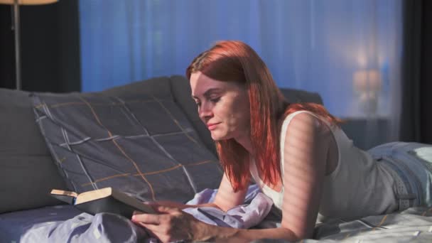 door te brengen tijd thuis, schattig jong vrouw lezen boek terwijl liggend op bed in de slaapkamer in zonneschijn - Video
