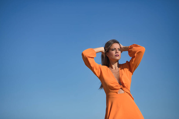 Jonge en mooie vrouw in een oranje jurk, met haar handen op haar hoofd, in sensuele houding, op het strand, met de wind door haar haar en jurk. Concept schoonheid, mode, trend, lucht. - Foto, afbeelding