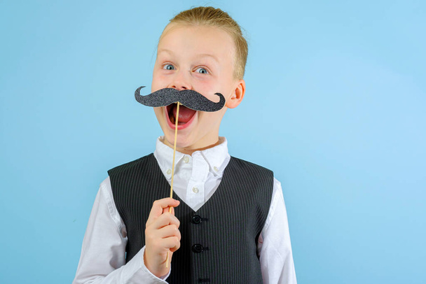 Funny child in an elegant suit holding mustache on a stick with his mouth wide open on the blue background. Purim celebration concept. Jewish carnival holiday. High quality photo - Photo, Image