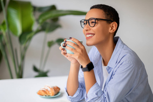 Счастливая спокойная молодая латинская женщина в очках наслаждается миром и обедом, пьет чашку кофе, ест круассан, смотрит на пустое пространство в современном кафе, офисный интерьер. Свободное время, отдых, завтрак - Фото, изображение