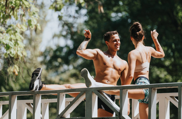 Ein Paar wärmt sich auf einer Brücke auf, dann trainiert es im Freien. Beharrlich fordern sie sich selbst heraus, um ihre Fitness-Ziele zu erreichen, umgeben von Freunden in einem Naturpark. - Foto, Bild