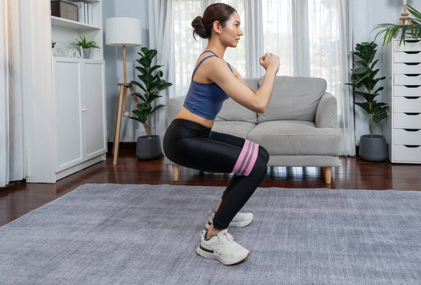 Silné energická žena dělá cvičení doma s odolnost sportovní pás pro svalové zisky nohou. Mladý atletický asijské žena síla a vytrvalost školení relace jako domácí cvičení rutina s squat. - Fotografie, Obrázek