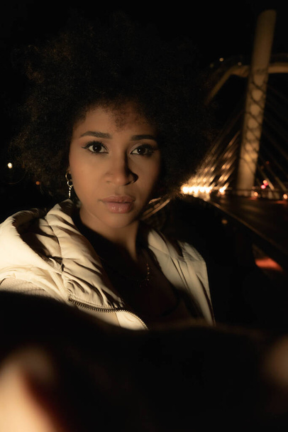 Афроамериканская женщина смотрит в камеру в городской обстановке ночью - Фото, изображение
